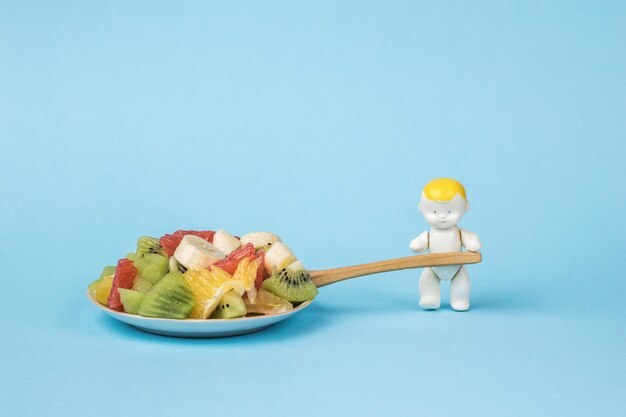 A baby figurine with a wooden spoon and a large bowl with fruit salad on a blue background.