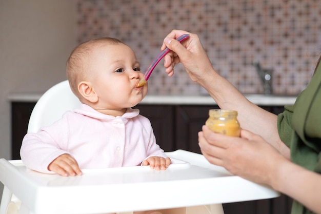 Baby food A baby in a high chair eats vegetable puree from a spoon Mom feeds the baby from a jar Kitchen Lifestyle