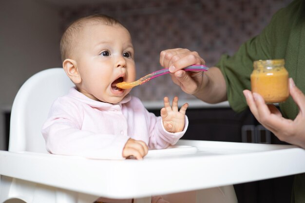 Baby food a baby in a high chair eats vegetable puree from a\
spoon mom feeds the baby from a jar kitchen lifestyle