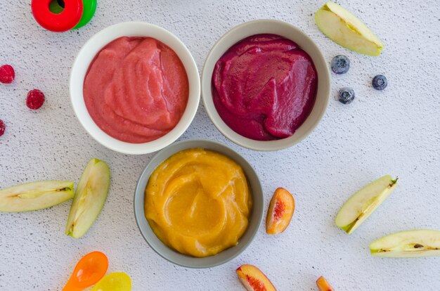 Baby food. Homemade fruit puree. Variety of apple puree or applesauce with frozen peach, raspberries and blueberries in three bowls on a light background.