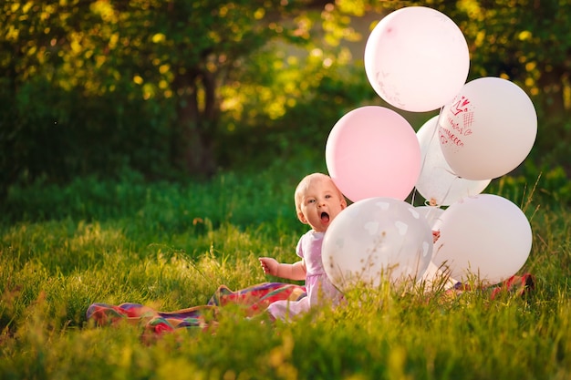 Baby girl 1 year old sitting on green grass with pink and white balloons in meadow outdoors closeup