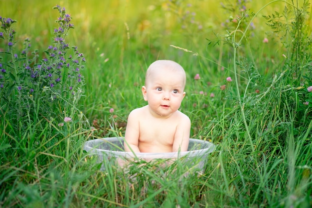 Baby girl 10 months old bathes in a basin in the grass in the summer.