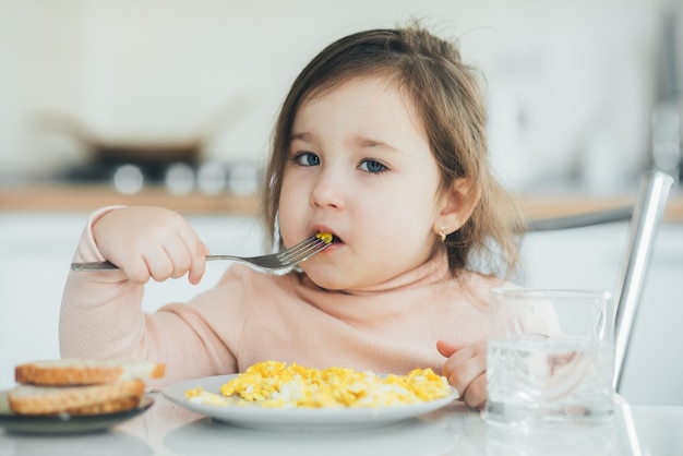 Bambina nel pomeriggio nella cucina a luce bianca con un maglione rosa che mangia una frittata