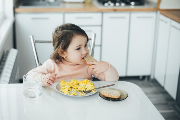 Baby girl in the afternoon in the white light kitchen in a pink sweater eating an omelet