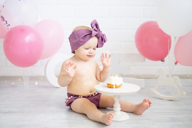 A baby girl celebrates her first year on her birthday with pink balloons and eats her cake with her hands smiles and rejoices