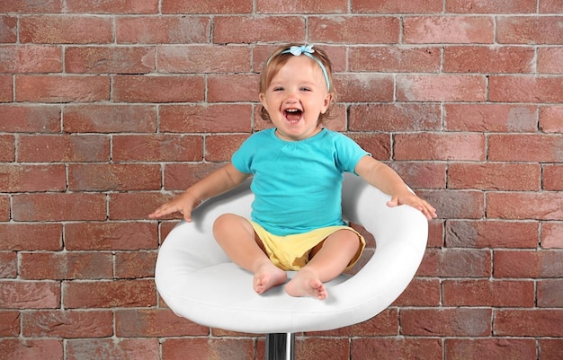 Baby girl in color dress sitting on a bar chair on a brick wall background