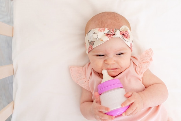 Baby girl does not want to eat a bottle of milk in the crib lying on her back in the nursery, baby food concept