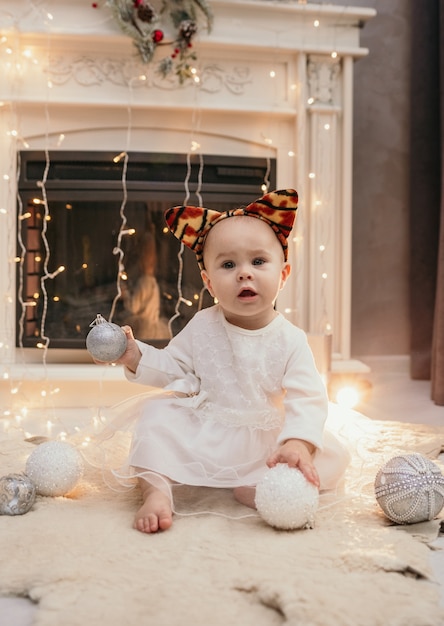 A baby girl in a fluffy dress and a headband with tiger ears sits near the fireplace in the room and plays with Christmas balls
