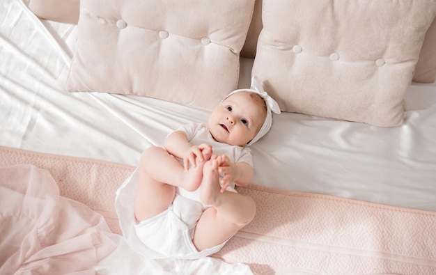 A baby girl in a headband and a white bodysuit is lying on the bed in the room