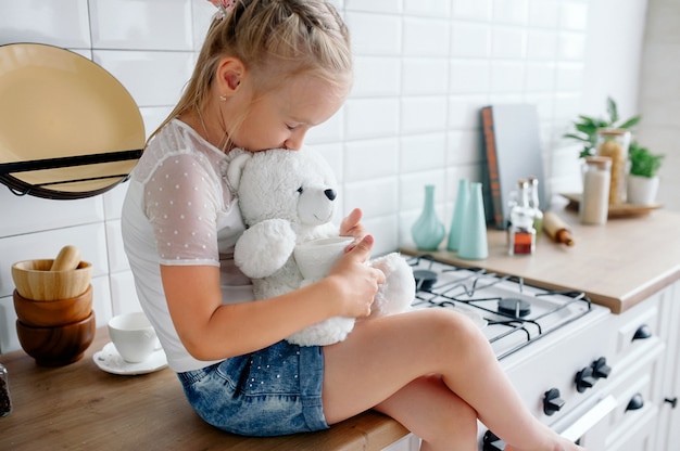 Baby girl hugs a teddy bear sitting in a beautiful stylish bright kitchen. Teddy Bear