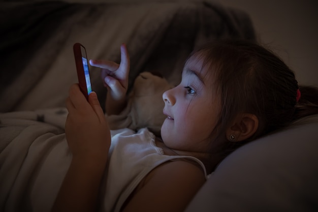 女の赤ちゃんがベッドに横になり、スマートフォンをクローズアップで使用します。漫画やゲームへの子供の中毒の概念。
