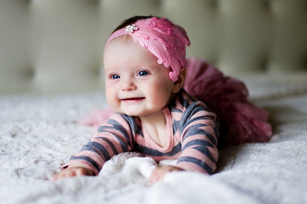 Photo baby girl lying on her stomach and smiles on the bed in clothes and a pink headband