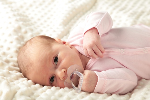 Baby girl lying on white bed and sucks a dummy, top view