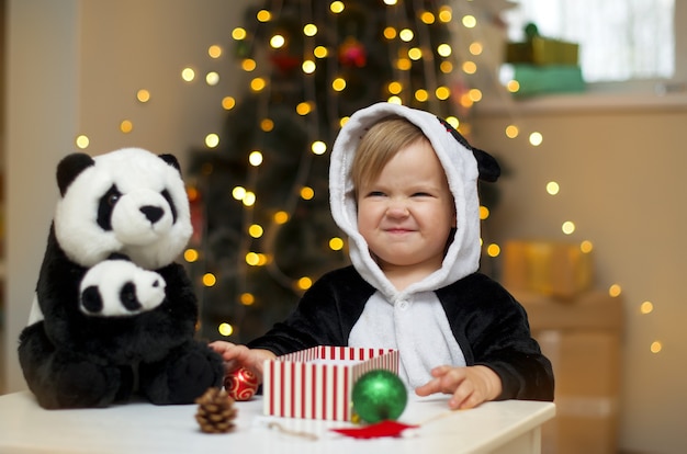 パンダのおもちゃでパンダの衣装を着た女の赤ちゃんがクリスマスツリーの下にクリスマスプレゼントを開きます