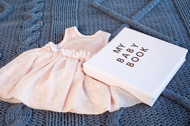 女の赤ちゃんピンクは、青い編み物の格子縞の背景に生まれたばかりの本のアルバムで服をドレスアップします。