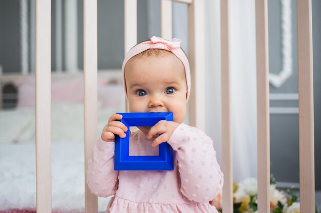 Baby girl in a pink dress sits and licks a toy in the crib. Teething