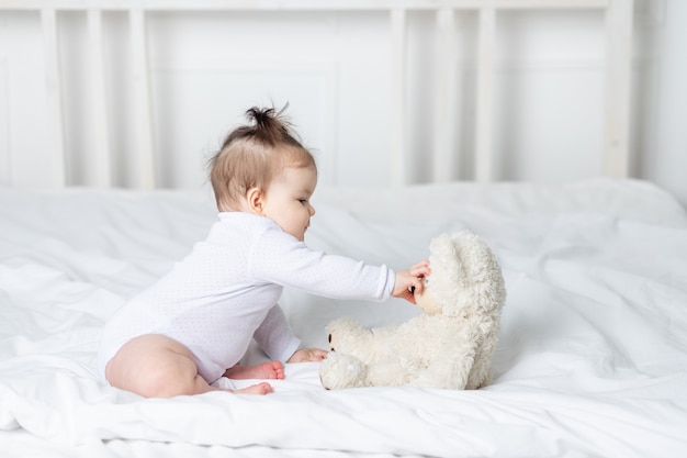 Baby girl playing with a teddy bear toy on the bed at home, the concept of play and development of children