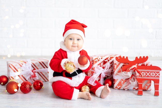 赤いサンタの衣装を着た女の赤ちゃんは、白い背景の上の贈り物やクリスマスのおもちゃの箱と一緒に座っています