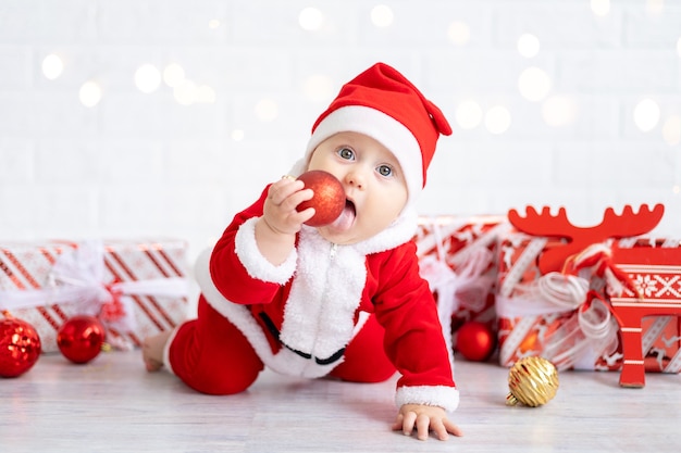 Baby girl in a red Santa costume sits with boxes of gifts and Christmas toys on a whitebackground