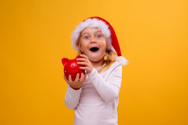 baby girl in Santa hat holding red piggy Bank on yellow wall, space for text