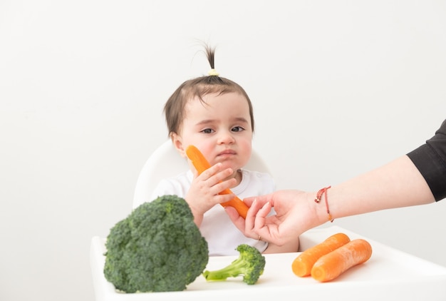 Baby girl sitting in baby chair eating carrot and broccoli.