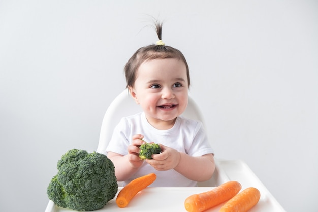 Baby girl sitting in baby chair eating carrot and broccoli.