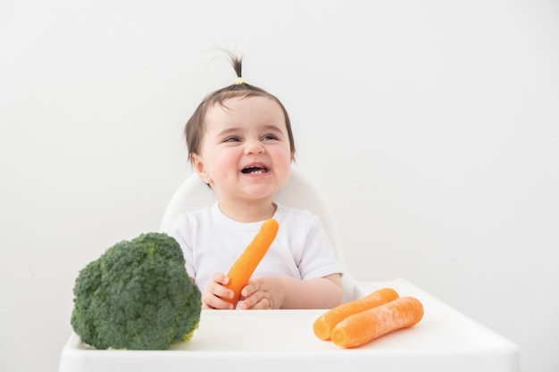 Baby girl sitting in baby chair eating carrot and broccoli.