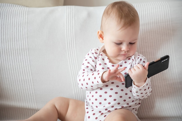 白いソファに座って黒いスマートフォンを持っている女の赤ちゃん