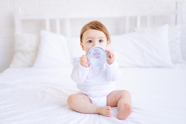 A baby girl of six months on a bed in white clothes sucks a bottle of water or milk a small child on a cotton bed at home woke up in the morning eating baby food concept