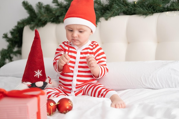 Baby girl in striped bodysuit and santa hat playing with christmas bell on decorated bed