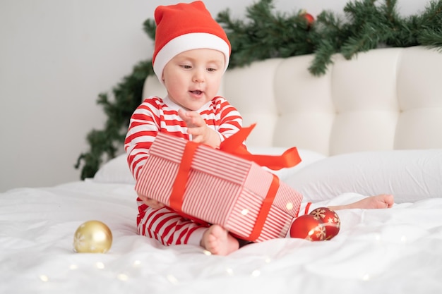 Baby girl in striped bodysuit and santa hat playing with gift box on christmas decorated bedroom