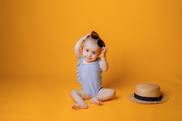 A baby girl in a swimsuit with a hat is sitting on a yellow background