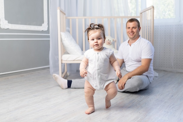 Baby girl takes her first steps, dad teaches baby to walk in the nursery, happy fatherhood