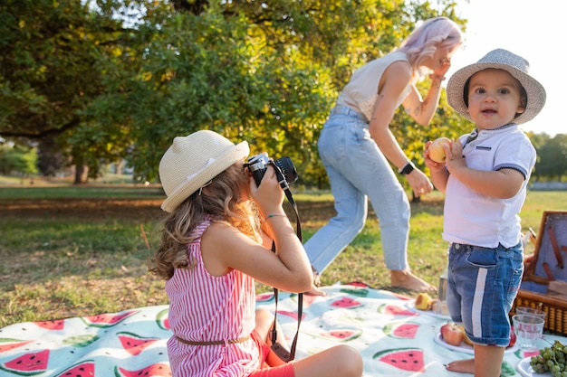 Baby girl takes photos of baby brother with analog camera while\
mom talks on phone during picnic