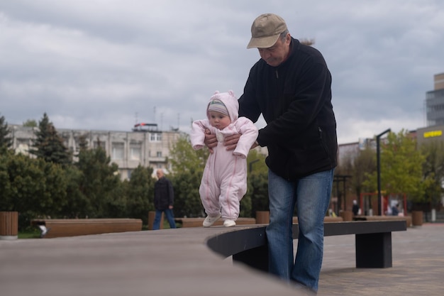 남자 할아버지와 함께 야외에서 걷는 아기 소녀는 그의 손녀가 걷도록 도와줍니다.