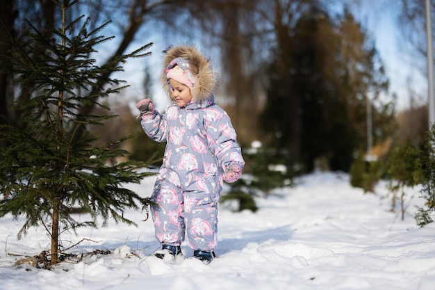사진 아기 소녀는 크리스마스 트리 근처의 맑은 서리가 내린 겨울날 아동 방한복을 입습니다.