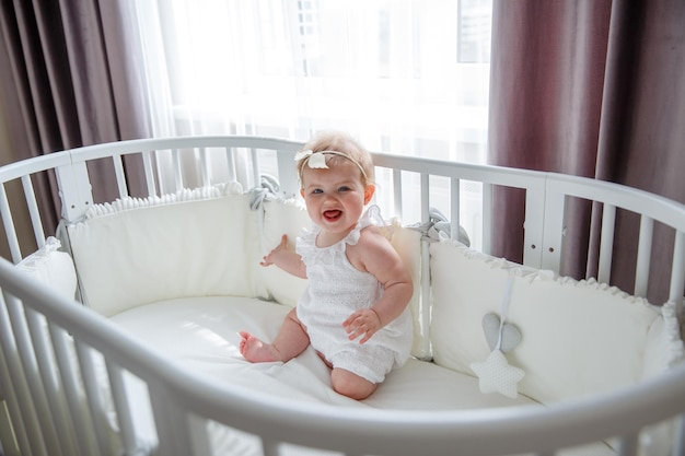 A baby girl in a white bodysuit is sitting in a crib in the\
bedroom