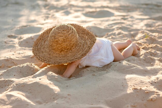 baby girl in white clothes and a straw hat sits on the white sand on the beach in summer