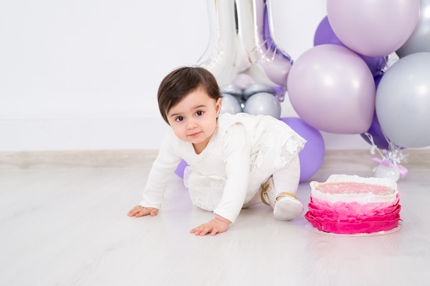 Baby girl in white dress sitting on the floor celebrating her first birthday with cake and balloons.