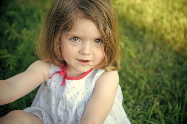 Baby girl with brown eyes on adorable face sitting on green grass on sunny summer day Innocence and youth concept Childhood leisure and fun