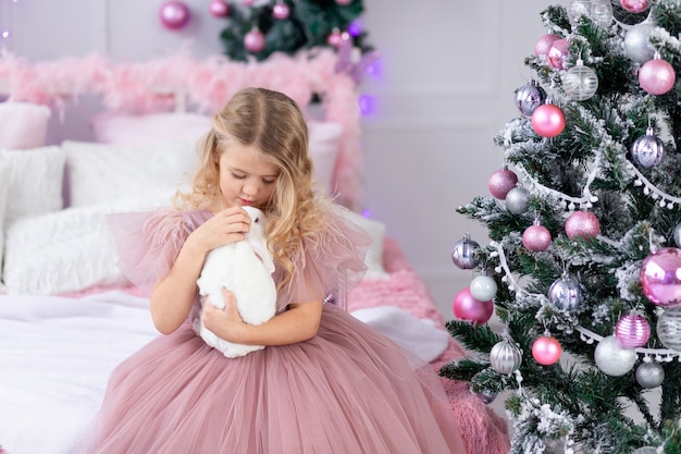 Baby girl with rabbit at the Christmas tree in a beautiful pink dress