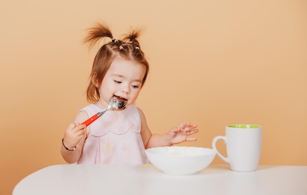 離乳食を食べるベージュ黄色のかわいい面白い赤ちゃんに分離されたスタジオでスプーンを持つ女の赤ちゃん