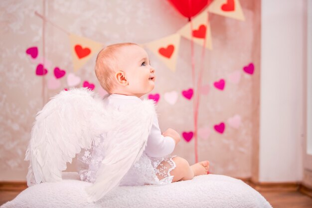 Baby girl with white feather wings sits on a white soft pillow among the Valentine's Day symbol, a view from the back.