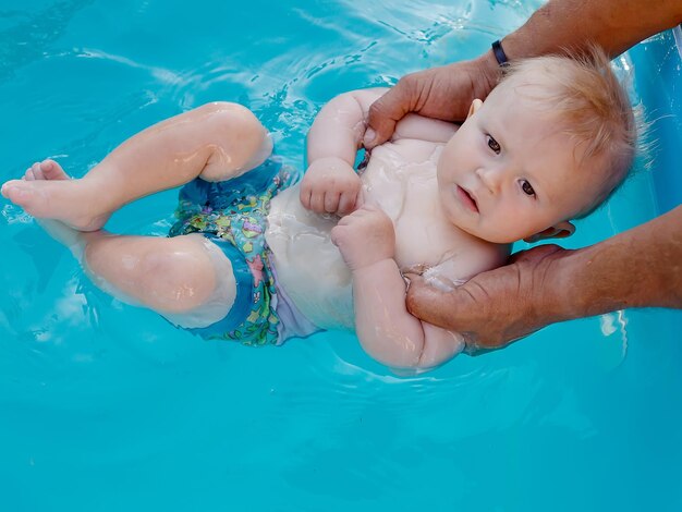 父の腕に抱かれた赤ちゃんが 足でプールの水を試みる