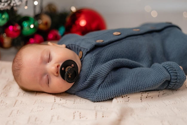 Baby in een blauwe gebreide jumpsuit met een zwarte fopspeen in zijn mond slaapt op een lichte deken op de vloer onder de kerstboom. eerste kerst. wintervakantie, grappige momenten. Hoge kwaliteit foto