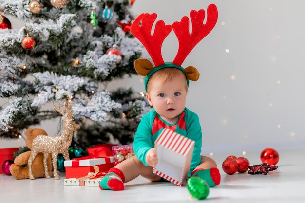 baby in een groen rompertje met edelhertenhoorns op zijn hoofd speelt naast de kerstboom