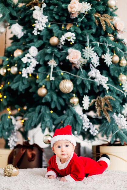 Baby in een kerstman kostuum liggend op een buik in de buurt van de kerstboom en cadeautjes