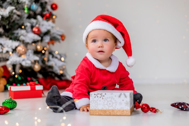 Baby in een kerstmankostuum zit naast de kerstboom en speelt met geschenkdozen. kind opent een kerstcadeau. winter nieuwjaarsconcept. ruimte voor tekst. hoge kwaliteit foto