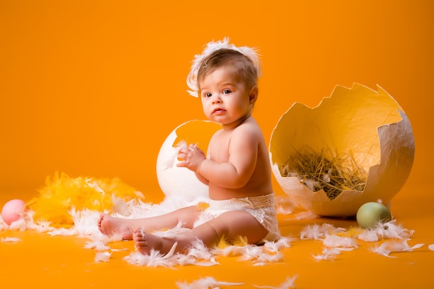 Baby in een kippenkostuum zit naast een decoratieve eierschaal op een gele achtergrond