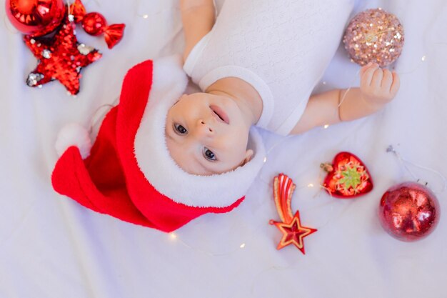 Baby in een witte bodysuit en een kerstmuts ligt op zijn rug op een wit laken omringd door rood kerstboomspeelgoed. winter, nieuwjaar. ruimte voor tekst. Hoge kwaliteit foto